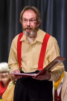 Man reading from a folder wearing a yellow shirt and red braces