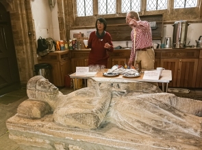 Two people, gesturing, behind a very old stone effigy